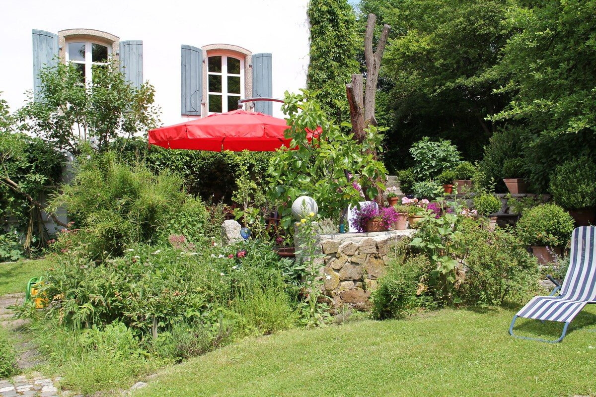 Sonniger Gartenplatz am Forsthaus mit rotem Sonnenschirm und altem Steinbrunnen.