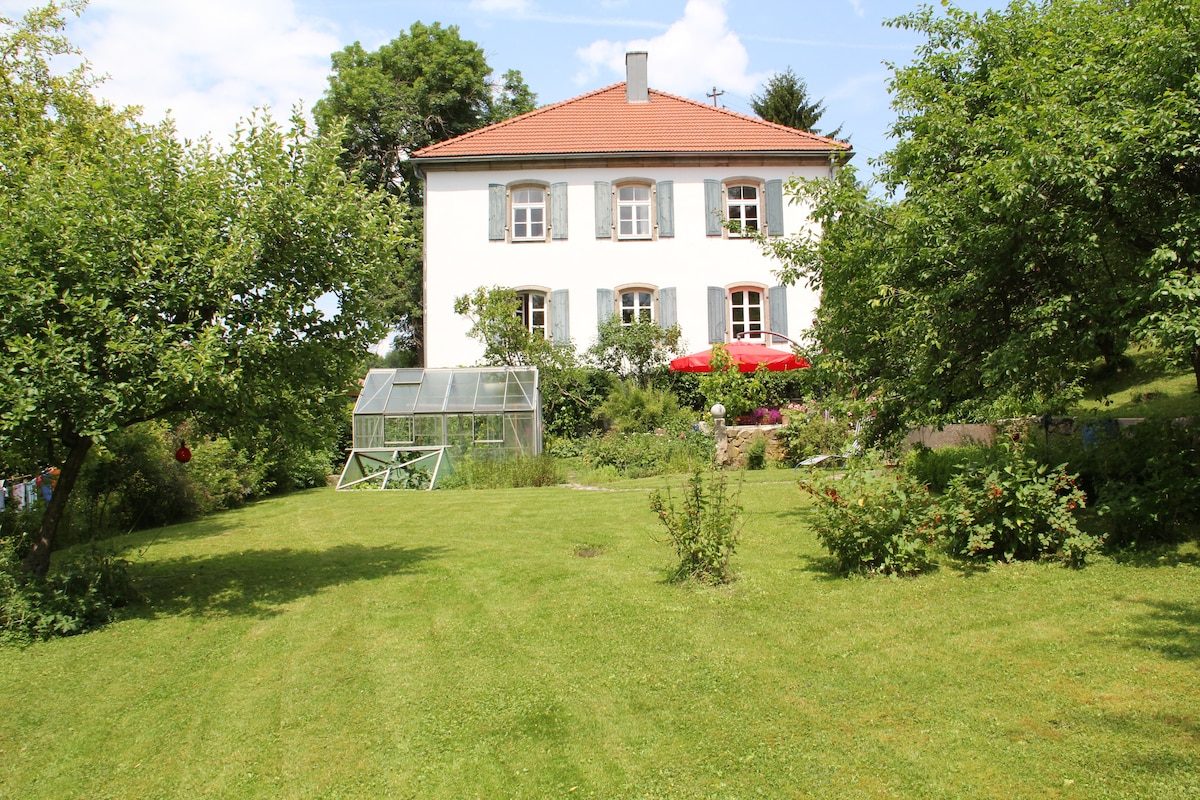 Das weiße Forsthaus von 1860 mit rotem Ziegeldach und blaugrauen Fensterläden, umgeben von einem weitläufigen grünen Garten mit Obstbäumen.