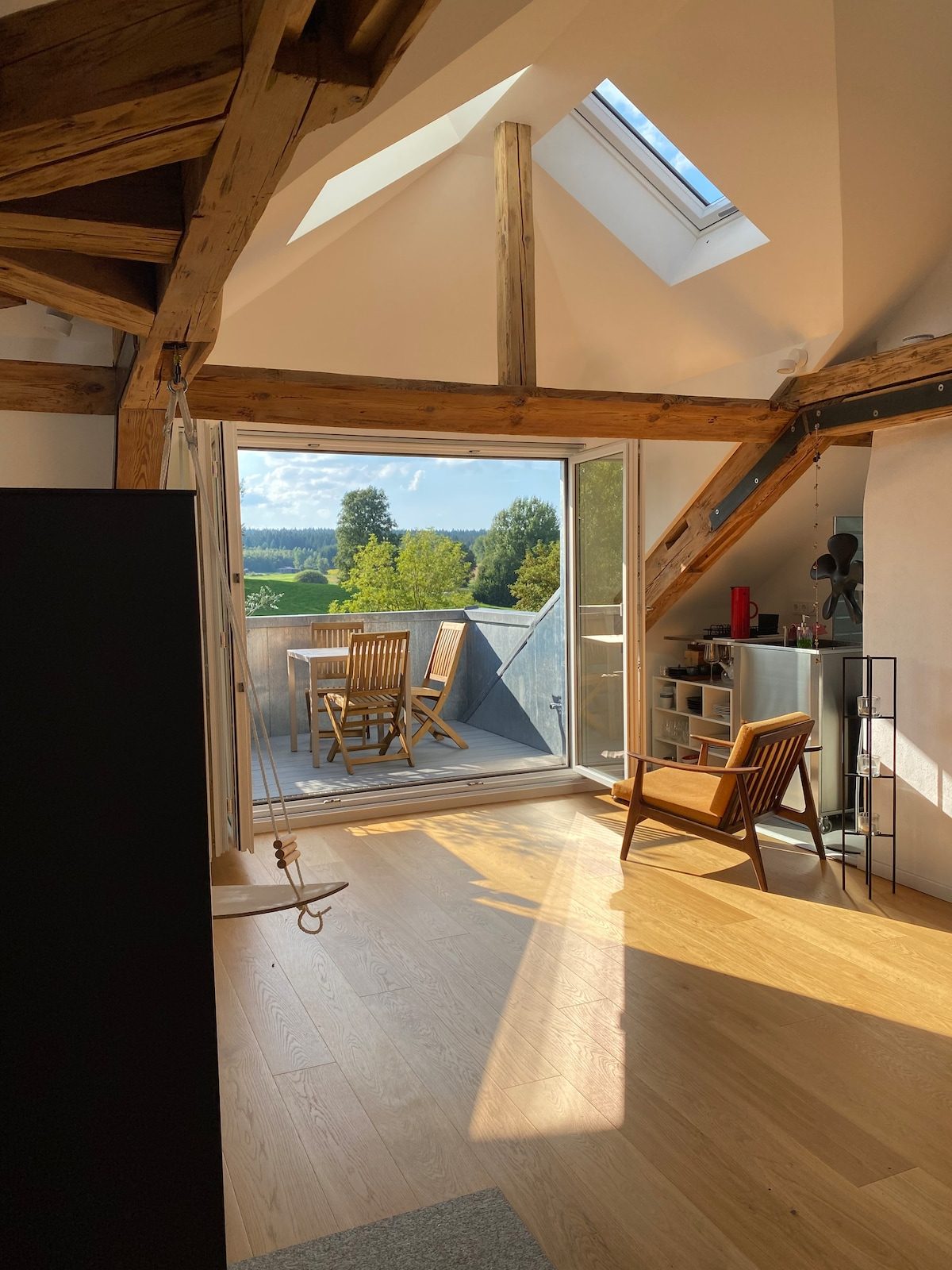 Loft-Wohnraum unter alten Dachbalken, bodentiefe Balkontür weit geöffnet mit Blick über Wiesen und Felder.